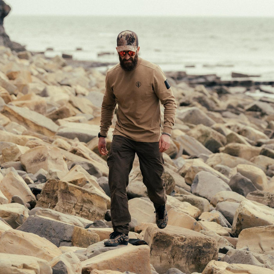 Louis Tinsley walking over rocks on the coast, wearing the Kryptek Cap with sunglasses, a Long Sleeve Insignia T-Shirt and the Foxy HeriTech Waxed Cotton Trousers.